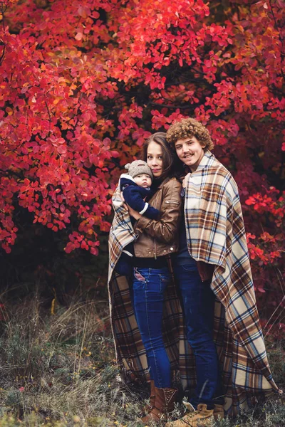 Familia feliz joven, cubierta de manta, se encuentra cerca de hermosos árboles rojos de otoño . — Foto de Stock