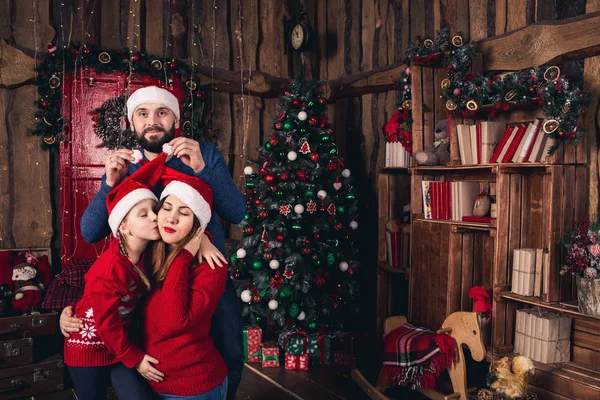 Happy family in Christmas hats sitting in a room in a rustic style. Father raises hats to his pregnant wife and daughter.