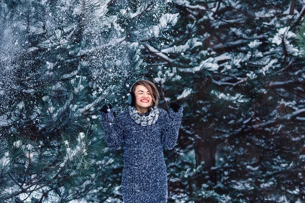 Stylish cheerful girl in a sweater in the winter forest. Snow from tree falls on the girl. — Stock Photo, Image