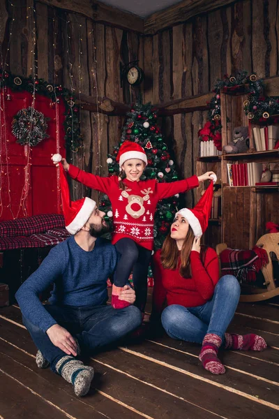 Gelukkige familie in Kerstmis hoeden zitten in een kamer in een rustieke stijl. Dochter verhoogt petje voor ouders. — Stockfoto