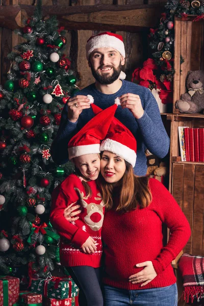 Gelukkige familie in Kerstmis hoeden zitten in een kamer in een rustieke stijl. Vader roept petje voor zijn zwangere vrouw en dochter. — Stockfoto