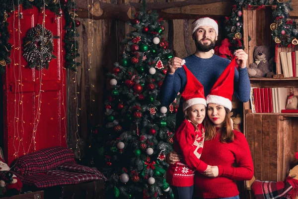 Gelukkige familie in Kerstmis hoeden zitten in een kamer in een rustieke stijl. Vader roept petje voor zijn zwangere vrouw en dochter. — Stockfoto