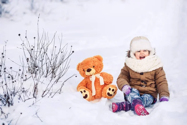 Une petite fille joyeuse est assise dans la neige avec son ours en peluche . — Photo