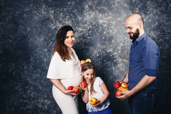Щаслива молода веганська сім'я. вагітна мати, бородатий батько, і маленька дочка тримає яблука в руках і посміхається — стокове фото