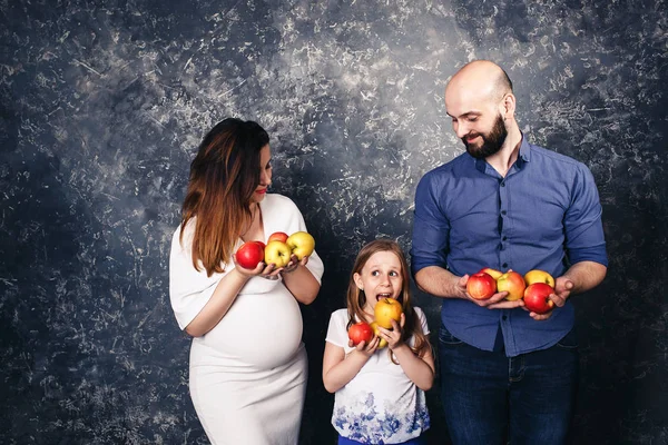 Вагітна мати, бородатий батько, і маленька дочка тримають яблука в руках і хочуть з'їсти їх — стокове фото