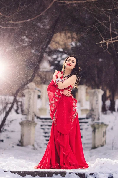 Krásná mladá dívka tančí břišní tance v červených šatech v zimě v parku na sněhu. — Stock fotografie
