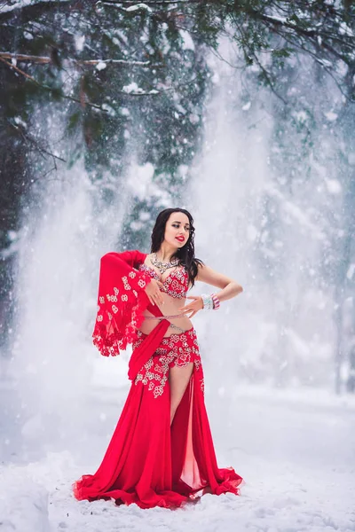 Beautiful young girl dancing belly dance in red dress in winter in a park on the snow. — Stock Photo, Image