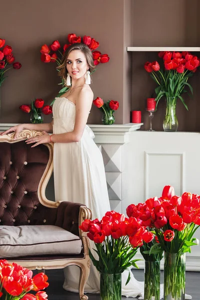 Hermosa joven en un vestido blanco posando en un estudio interior decorado con grandes ramos de tulipanes rojos. 8 de marzo concepto. Mañana de la novia . — Foto de Stock