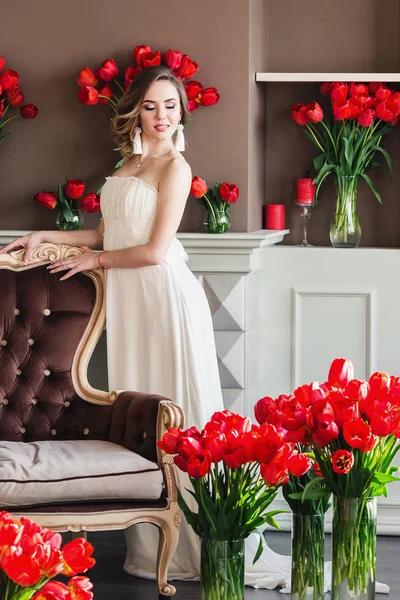 Hermosa joven en un vestido blanco posando en un estudio interior decorado con grandes ramos de tulipanes rojos. 8 de marzo concepto. Mañana de la novia . — Foto de Stock