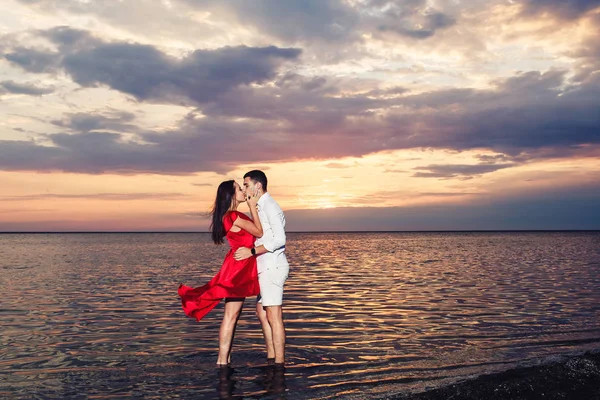 Joven pareja enamorada besos de pie en el mar sobre el telón de fondo de una hermosa puesta de sol . —  Fotos de Stock