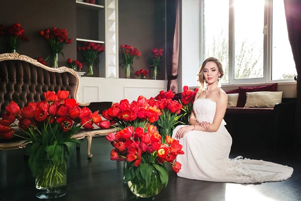 Hermosa joven en un vestido blanco posando en un estudio interior decorado con grandes ramos de tulipanes rojos. 8 de marzo concepto. Mañana de la novia . — Foto de Stock