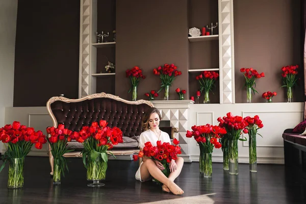 Una hermosa joven en un peignoir está sentada en el suelo en la sala de estar entre grandes ramos de tulipanes. Concepto 8 de marzo . — Foto de Stock