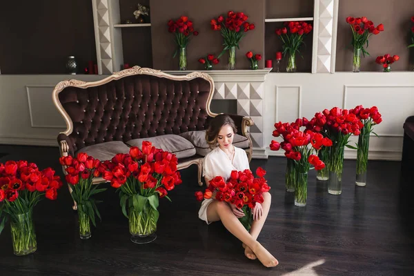 Krásná mladá dívka v peignoir sedí na podlaze v obývacím pokoji mezi velké Kytice tulipánů. 8. března koncepce. — Stock fotografie