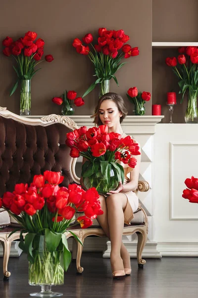 Una hermosa joven con un vestido blanco se sienta en el sofá y sostiene un gran ramo de tulipanes rojos. 8 de marzo concepto. Mañana de la novia . — Foto de Stock