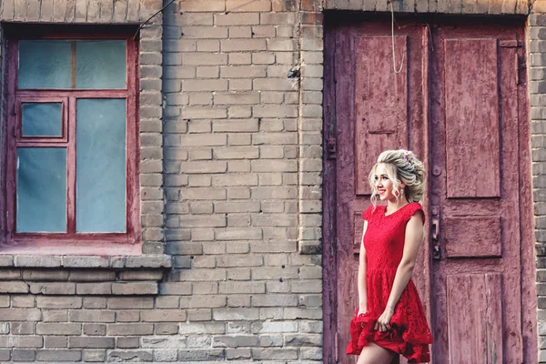 Loira jovem atraente em vestido vermelho posando no fundo de um edifício de tijolo velho . — Fotografia de Stock