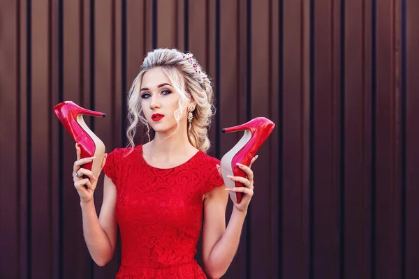 Atractiva joven rubia en un vestido rojo sostiene en las manos zapatos rojos . — Foto de Stock