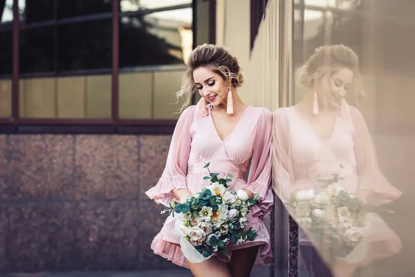 Chica joven atractiva en un vestido corto con un ramo de flores posando en la calle cerca de la pared del edificio . — Foto de Stock