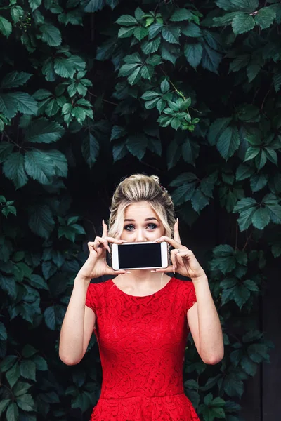Atractiva rubia joven en un vestido rojo cierra la boca con un teléfono inteligente . — Foto de Stock