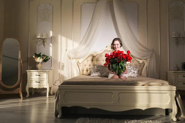 Una hermosa joven está sentada en la cama y sosteniendo muchos tulipanes rojos en un jarrón. 8 de marzo concepto . — Foto de Stock