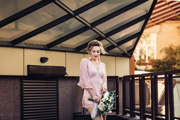 Jovem atraente em um vestido curto com um buquê de flores posando na rua perto da parede do edifício . — Fotografia de Stock