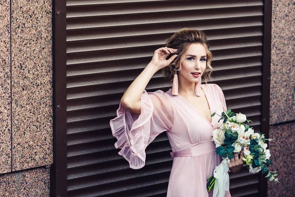 Jovem atraente em um vestido curto com um buquê de flores posando perto da parede de granito com persianas . — Fotografia de Stock