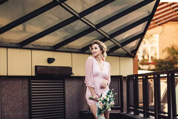 Jovem atraente em um vestido curto com um buquê de flores posando na rua perto da parede do edifício . — Fotografia de Stock