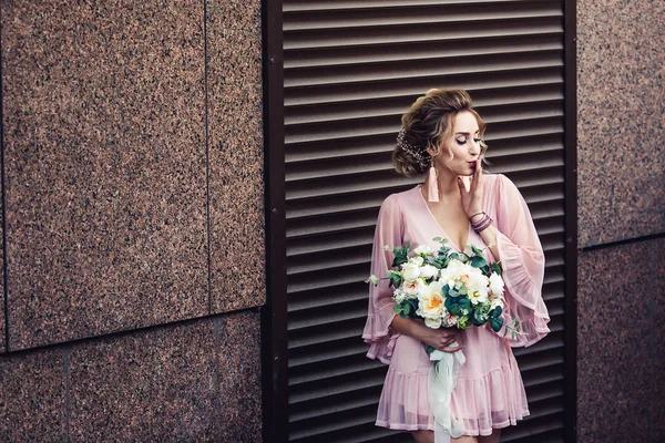 Attraktive junge Mädchen in einem kurzen Kleid mit einem Blumenstrauß posiert in der Nähe der Granitwand mit Rollläden. — Stockfoto