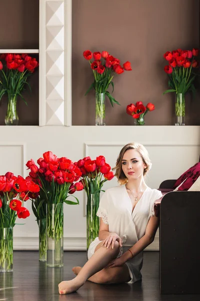 Una hermosa joven en un peignoir está sentada en el suelo en el estudio con ramos de tulipanes. El concepto del 8 de marzo. Mañana de la novia . — Foto de Stock