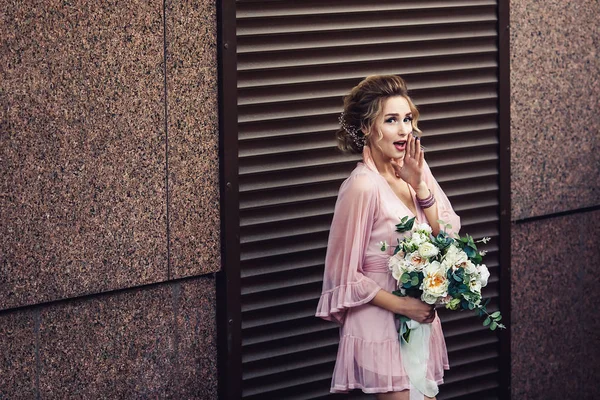 Jovem atraente em um vestido curto com um buquê de flores posando perto da parede de granito com persianas . — Fotografia de Stock