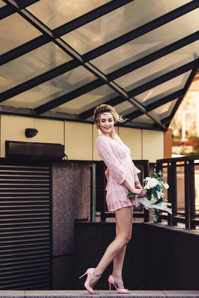 Jovem atraente em um vestido curto com um buquê de flores posando na rua perto da parede do edifício . — Fotografia de Stock