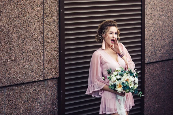 Jovem atraente em um vestido curto com um buquê de flores posando perto da parede de granito com persianas . — Fotografia de Stock