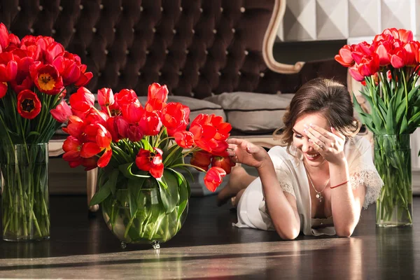 Una hermosa joven en un peignoir está tirada en el suelo en el estudio con ramos de tulipanes. El concepto del 8 de marzo. Mañana de la novia . — Foto de Stock