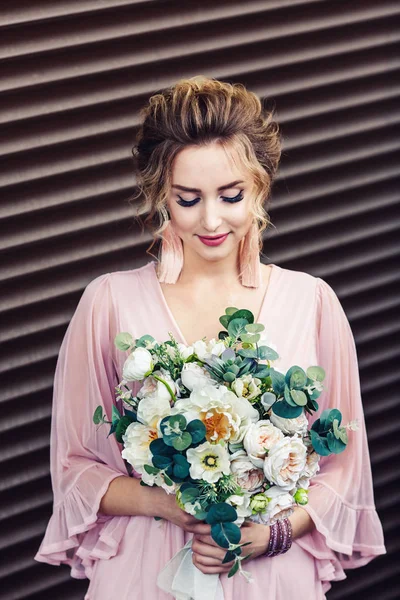 Jovem atraente em um vestido curto com um buquê de flores posando perto da parede de granito com persianas . — Fotografia de Stock