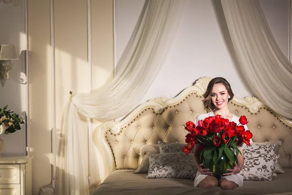 Una hermosa joven está sentada en la cama y sosteniendo muchos tulipanes rojos en un jarrón. 8 de marzo concepto . — Foto de Stock