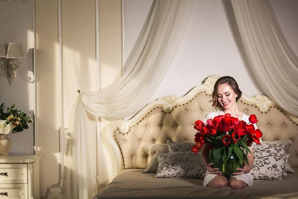 Una hermosa joven está sentada en la cama y sosteniendo muchos tulipanes rojos en un jarrón. 8 de marzo concepto . — Foto de Stock