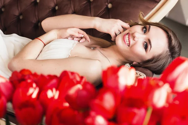 Una hermosa joven está tumbada en el sofá, y frente a ella hay tulipanes rojos. 8 de marzo concepto. Mañana de la novia . — Foto de Stock