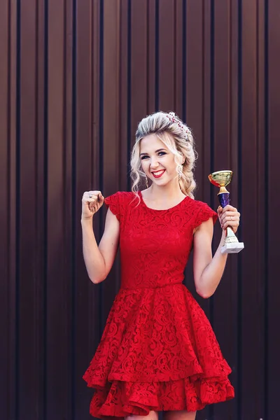 Atractiva rubia joven con un vestido rojo sosteniendo la copa de un ganador en sus manos sobre un fondo marrón . — Foto de Stock