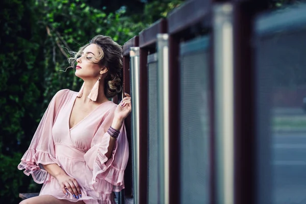 Atractiva joven en un vestido corto de color rosa posando cerca de una valla de acero en el fondo de follaje verde . — Foto de Stock