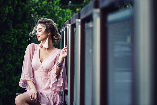 Atractiva joven en un vestido corto de color rosa posando cerca de una valla de acero en el fondo de follaje verde . — Foto de Stock