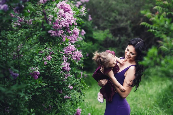แม่และลูกสาวที่มีความสุขในชุดสีม่วง เล่นในธรรมชาติใกล้ดอกลิลลาค — ภาพถ่ายสต็อก