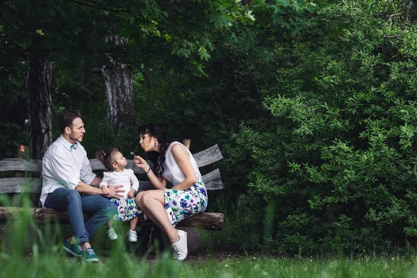 Friendly happy family resting on a wooden bench and playing with a dandelion — Stock Photo, Image