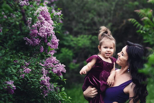 Madre alegre e hija pequeña en vestidos púrpura jugando en la naturaleza cerca de la floreciente lila — Foto de Stock