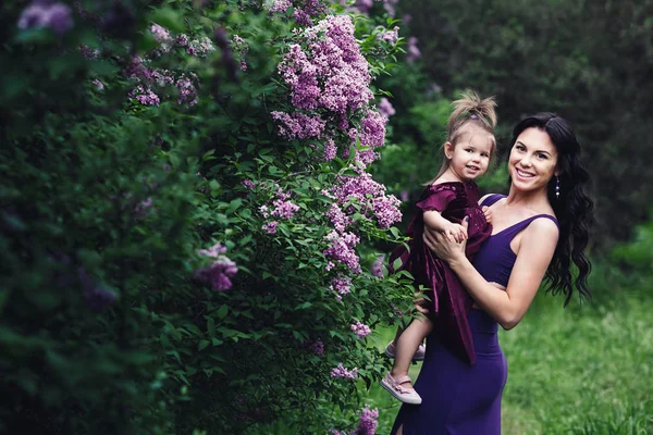 Madre alegre e hija pequeña en vestidos púrpura jugando en la naturaleza cerca de la floreciente lila — Foto de Stock