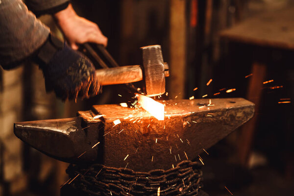 The blacksmith manually forging the red-hot metal on the anvil in smithy with spark fireworks.