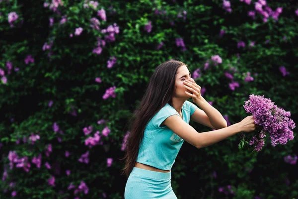 Seizoengebonden allergieën. Een jong meisje niest in een park te midden van lila bloemen en in het bezit van een boeket. — Stockfoto