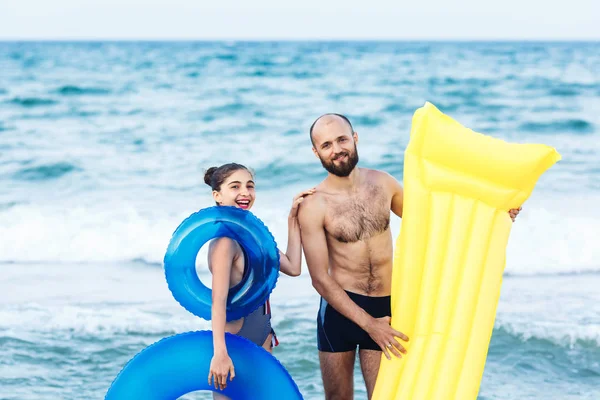 Radosny ojciec i córka stoją na plaży trzymając dmuchany materac i okrąg. — Zdjęcie stockowe