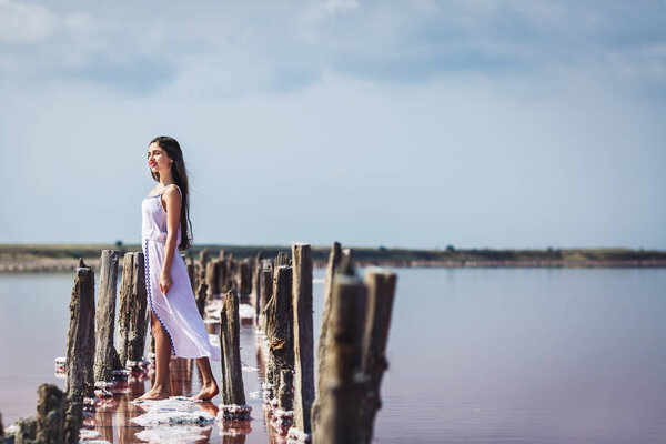 beautiful young girl in long white dress posing on salty pink lake.