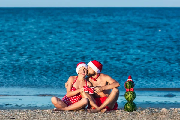 Brodaty mężczyzna i dziewczynka w Santa ubrania całują się na plaży morskiej z kubków w rękach i bałwana z arbuz. Letnie święta Bożego Narodzenia. — Zdjęcie stockowe