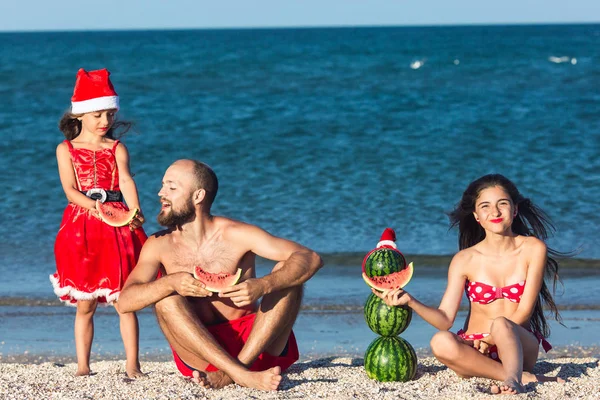Merry family celebrates summer Christmas. father and two daughters eating watermelon on beach.