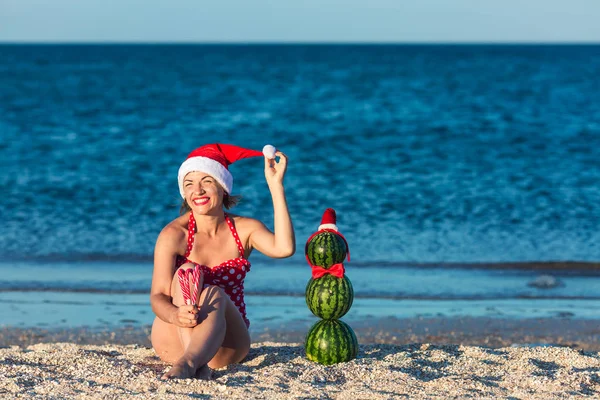 Radosna młoda kobieta w kapeluszu Mikołaja trzyma laski świąteczne cukierki, i arbuz bałwana stoi w pobliżu. — Zdjęcie stockowe